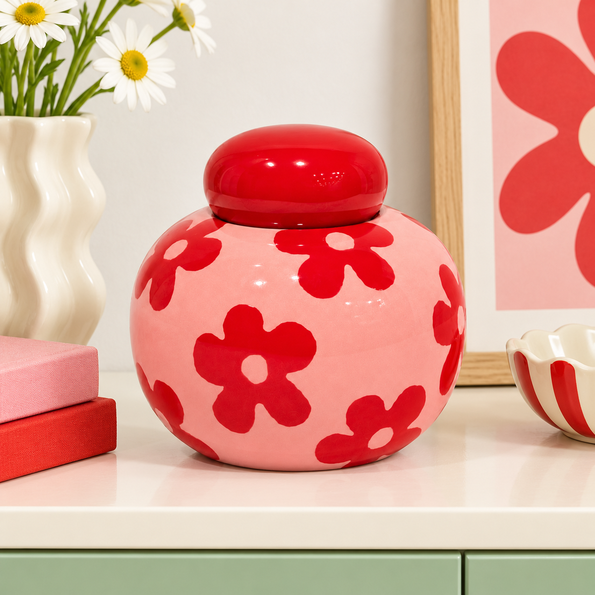 Barattolo Simona Fiori di Qué Rico rosa e rosso, ambientato su un mobile chiaro accanto a un vaso con margherite e complementi decorativi.