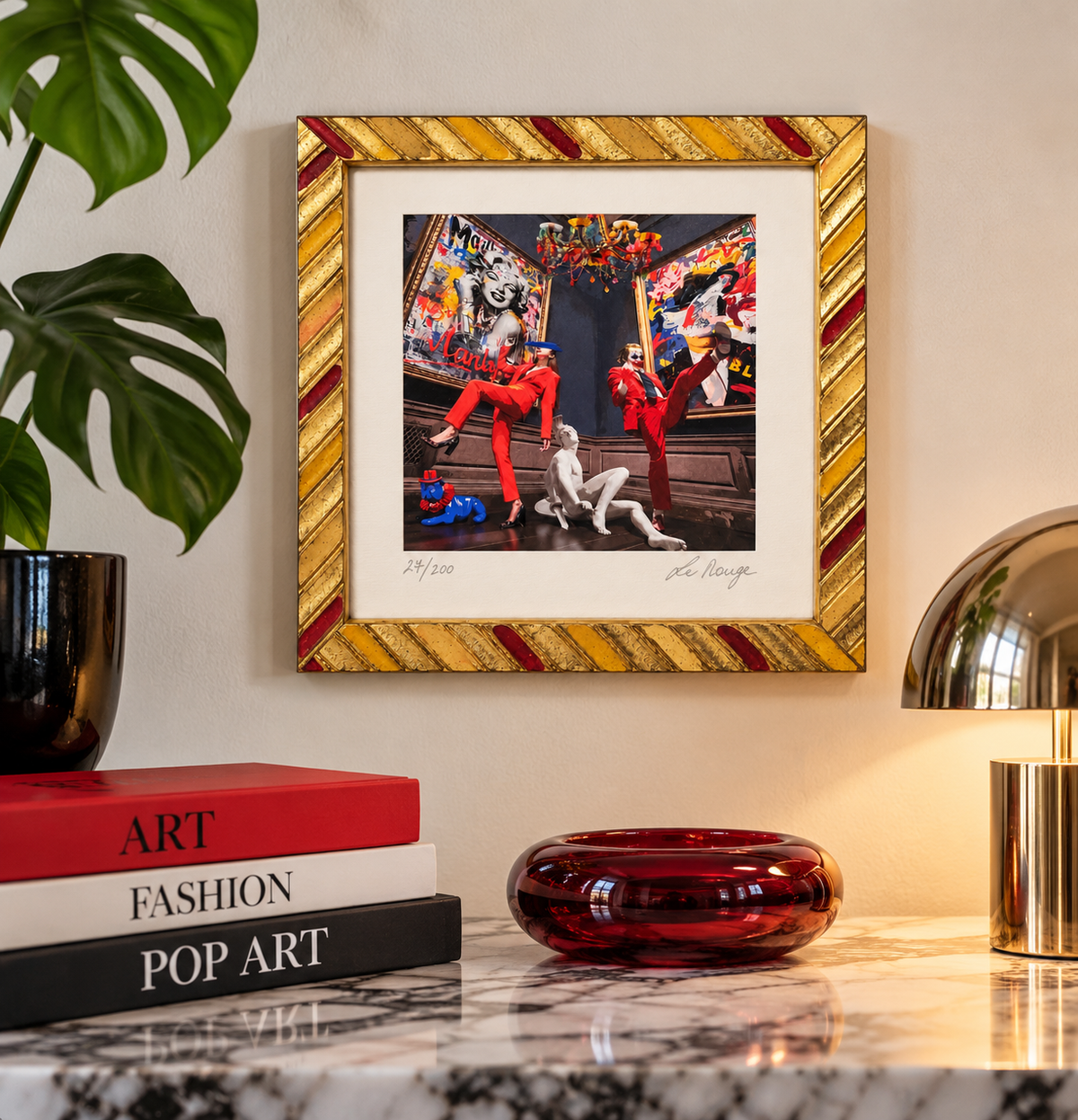 Serigrafia Fashion Street di La Rouge con cornice dorata e rossa, esposta su una parete chiara sopra una consolle in marmo con libri d'arte e lampada dorata.