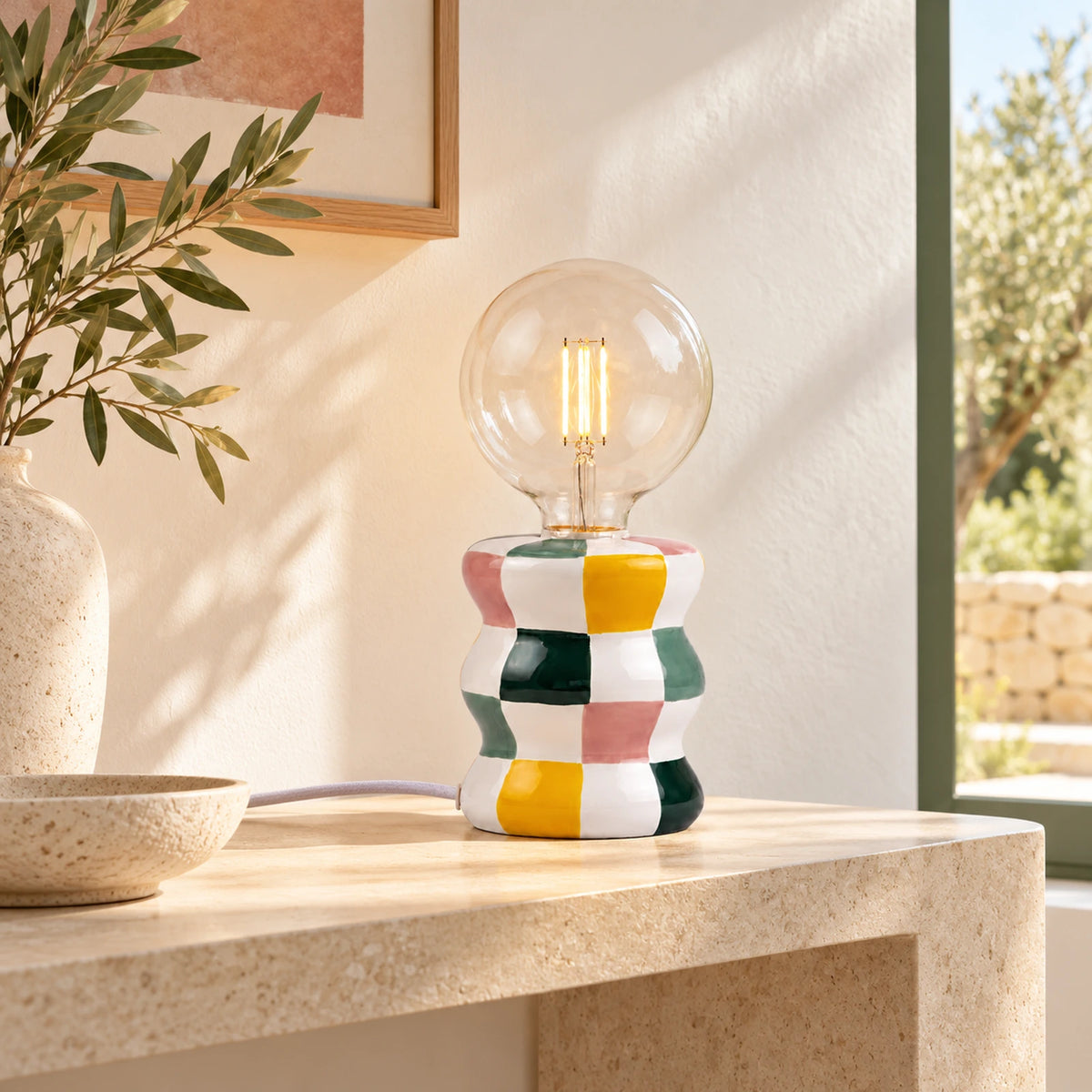 Lampada Ektor Qué Rico accesa, con base in ceramica a scacchi multicolore e lampadina sferica, ambientata su una consolle in pietra.