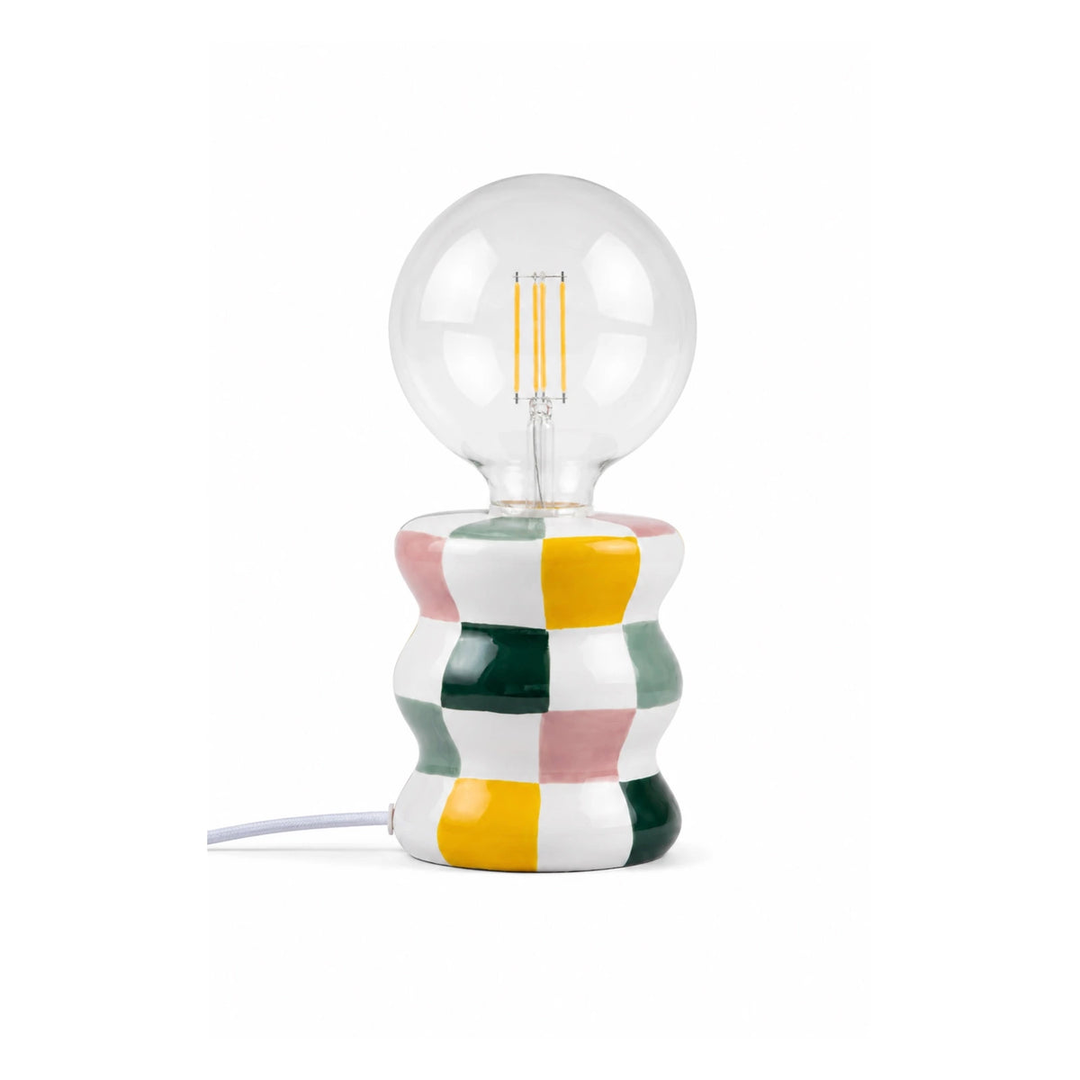 Lampada da tavolo Ektor di Qué Rico con base in ceramica a scacchi rosa, gialli, verdi e bianchi e lampadina sferica a vista, su fondo bianco.