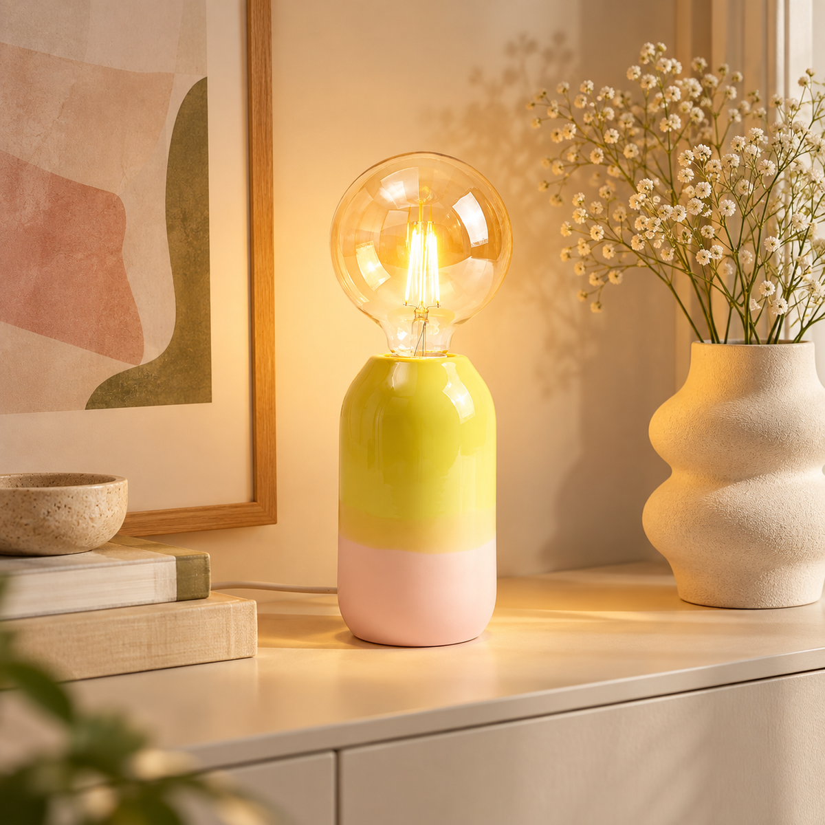 Lampada Farol Motel a Miio giallo lime e rosa, accesa su una consolle chiara accanto a libri, stampa astratta e vaso con fiori.