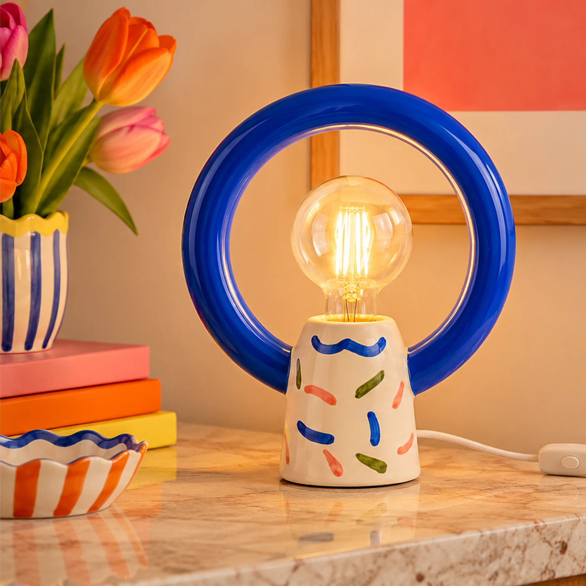 Lampada Matias Joyful Jumble Qué Rico accesa, con struttura circolare blu e base multicolore, ambientata su una consolle con libri e fiori.