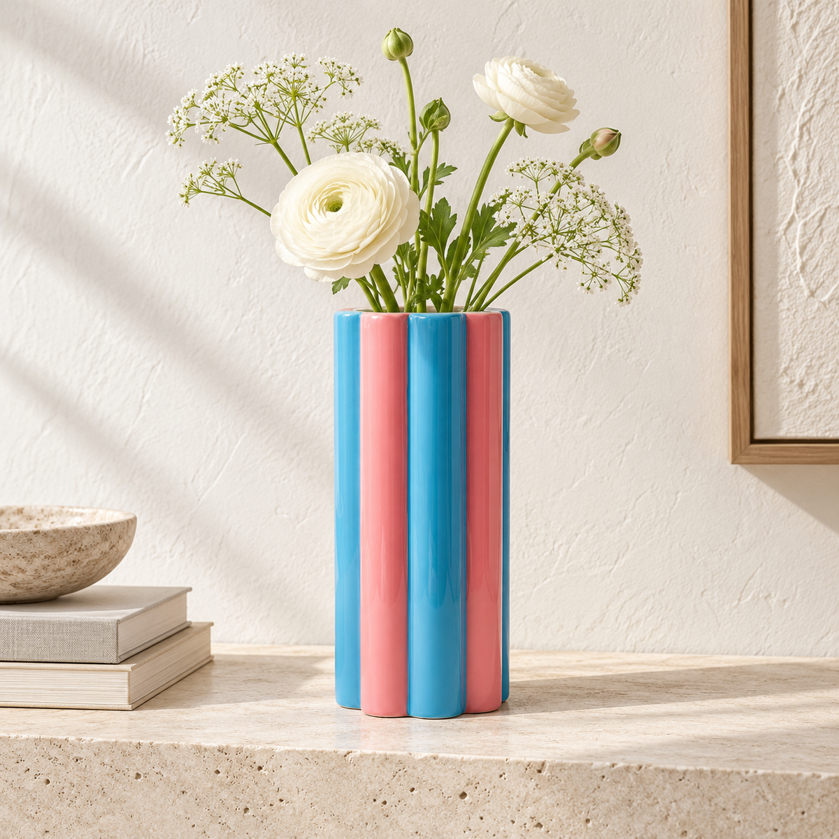 Vaso Arturo Qué Rico azzurro e rosa con fiori bianchi delicati, ambientato su un piano in pietra con libri decorativi.
