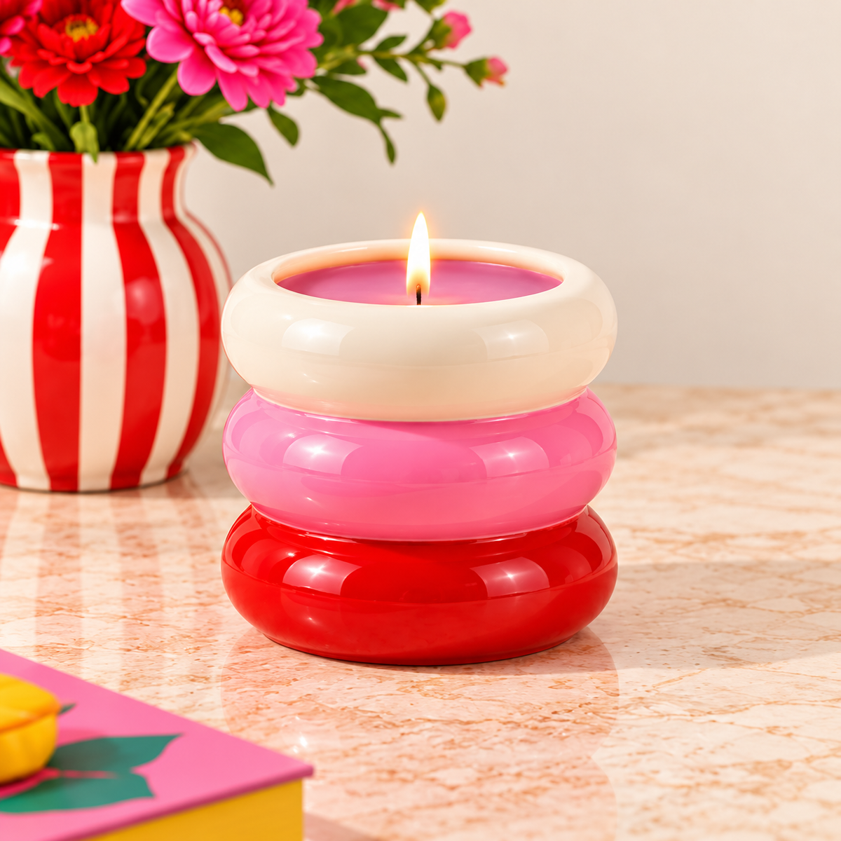 Candela Qué Rico in vaso ad anelli bianchi, rosa e rossi, accesa su un mobile con fiori e complementi decorativi.
