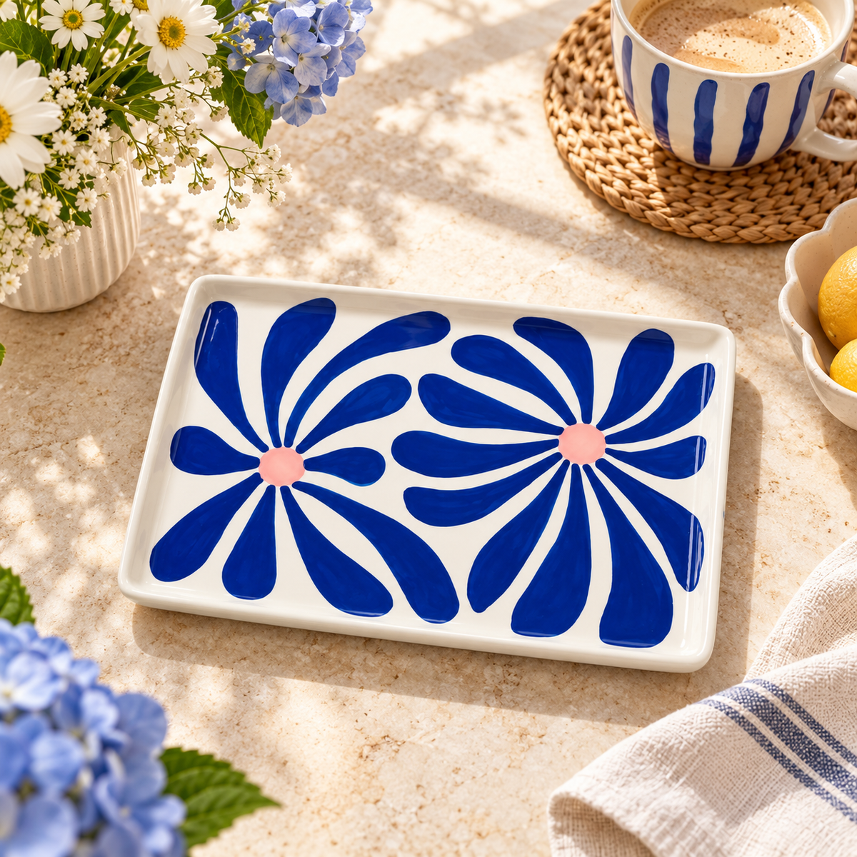 Vassoio Alejandro Qué Rico con grandi fiori blu e centri rosa, ambientato su un piano in pietra chiara con margherite, fiori azzurri e tessuto naturale.