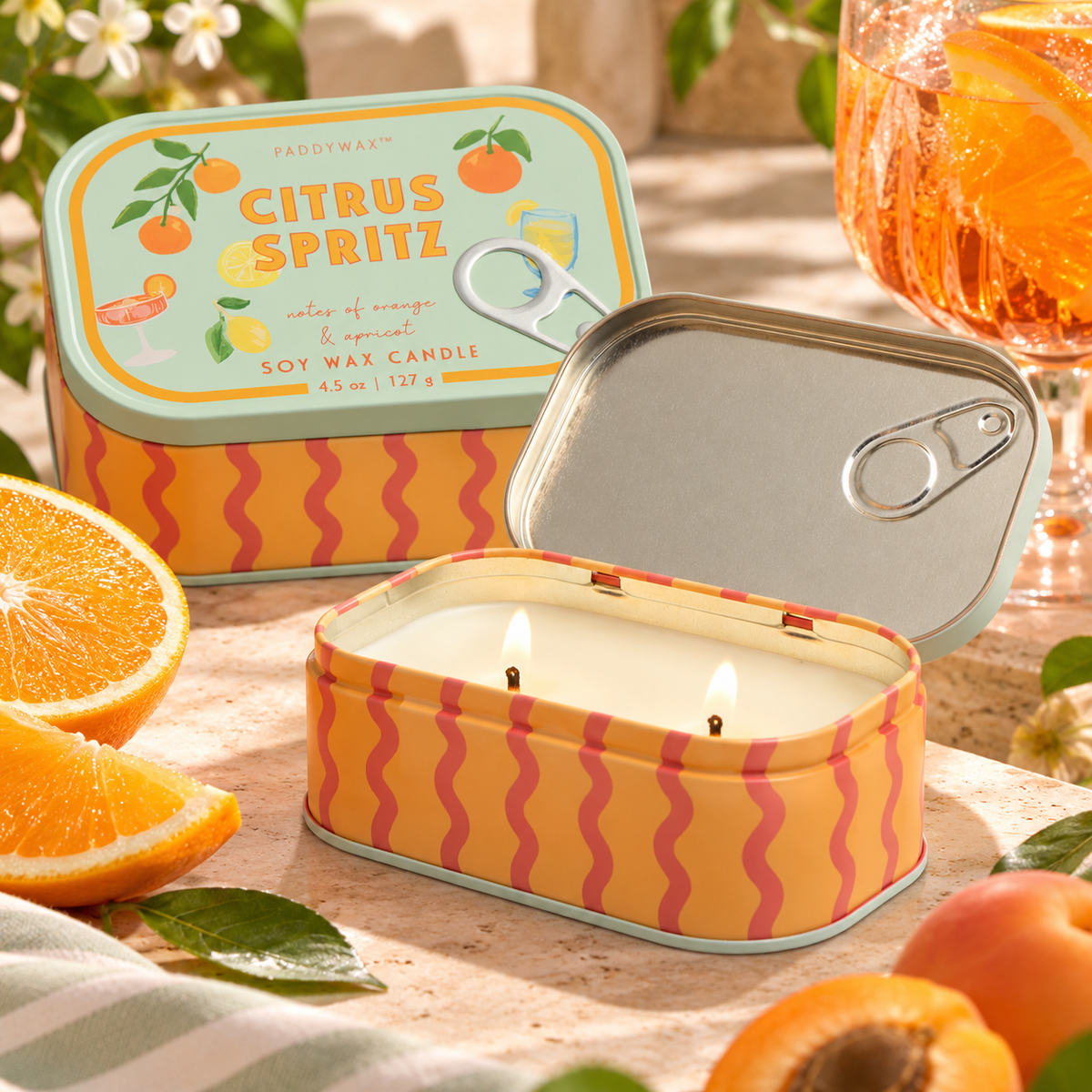 Candela Paddywax Citrus Spritz aperta e accesa con due stoppini, in lattina arancione decorata, ambientata con agrumi e un cocktail.