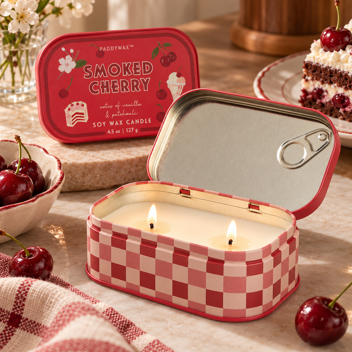 Candela Paddywax Smoked Cherry aperta e accesa con due stoppini, in lattina a quadretti rossi e rosa, ambientata accanto a ciliegie e dolci.