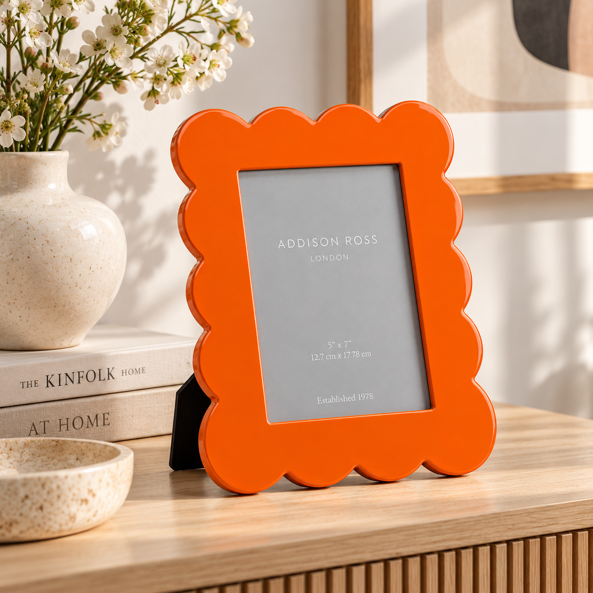 Cornice sagomata Addison Ross arancione laccata lucida, ambientata su una consolle in legno accanto a libri e vaso con fiori bianchi.