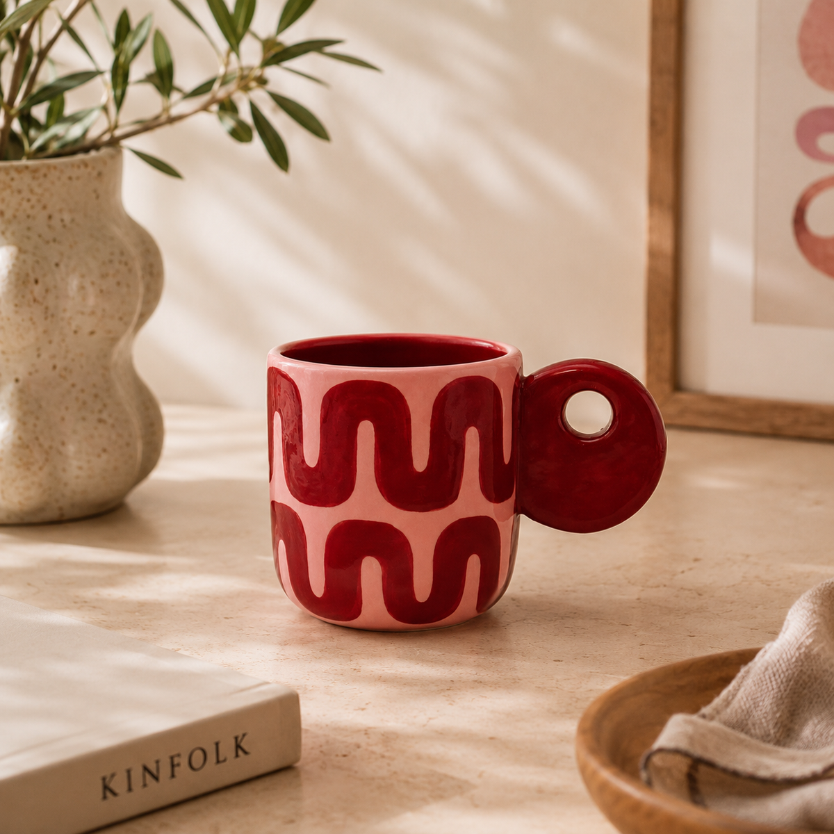 Mug Leonor Qué Rico rosa con motivi ondulati bordeaux e manico circolare, ambientata su un piano in pietra chiara accanto a un libro Kinfolk.