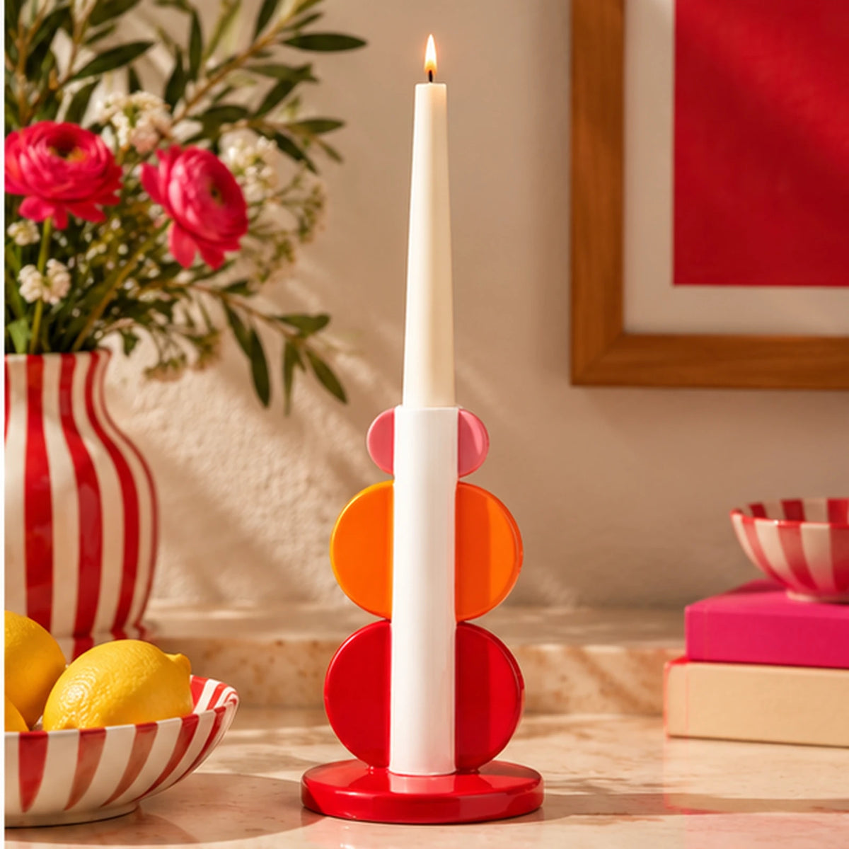 Portacandele Lorenzo Qué Rico rosso, arancione e rosa con lunga candela bianca accesa, ambientato su una consolle in pietra con fiori decorativi.
