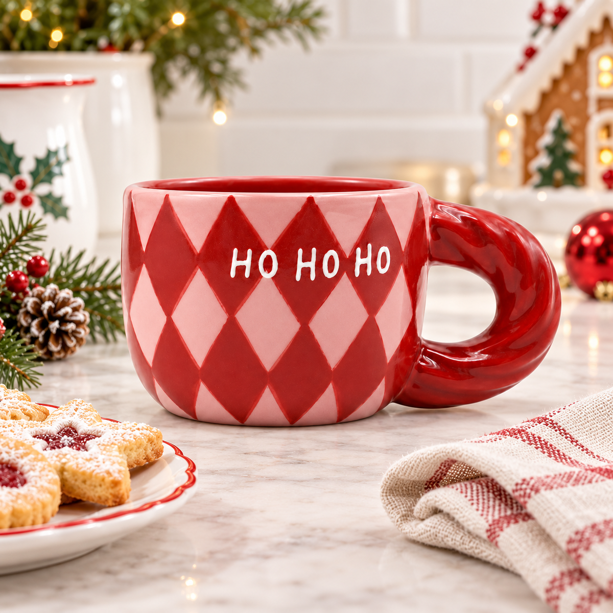 Tazza natalizia Natalia Qué Rico rosa e bordeaux con scritta HO HO HO, ambientata su un tavolo chiaro con biscotti, decorazioni e luci di Natale.