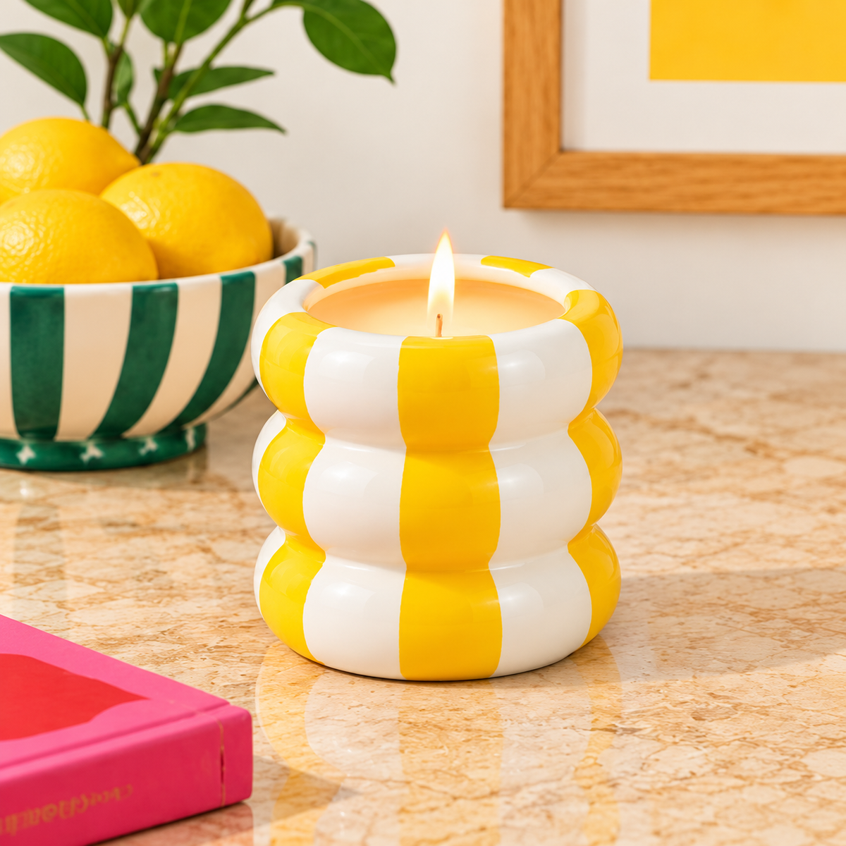 Candela Qué Rico in vaso giallo e bianco a fasce arrotondate, accesa su un piano in pietra con limoni e oggetti decorativi.