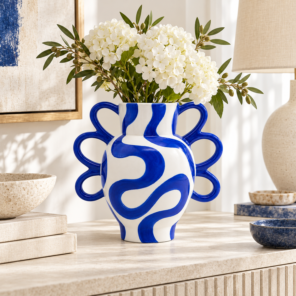 Vaso Yago Crazy Curves Qué Rico bianco e blu cobalto con fiori bianchi, ambientato su una consolle in pietra chiara in un soggiorno contemporaneo.